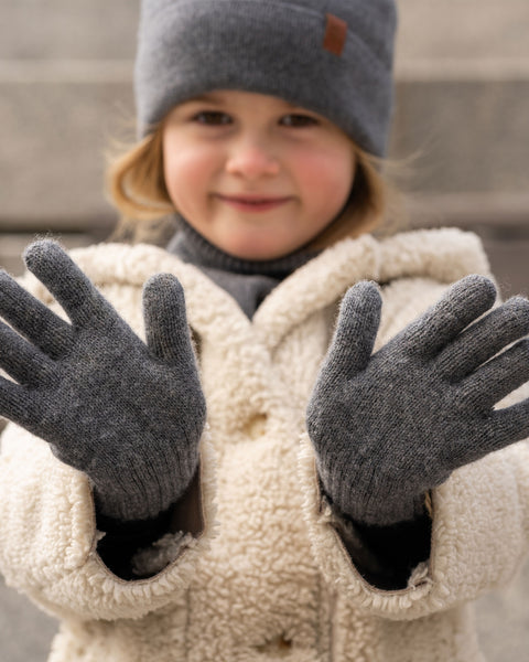 A gently smiling young child holding both hands out towards the camera, wearing dark grey knitted gloves. The child is dressed in a matching dark grey beanie, a dark grey neck warmer, and a fluffy cream-colored fleece jacket. The background features softly blurred stone steps.