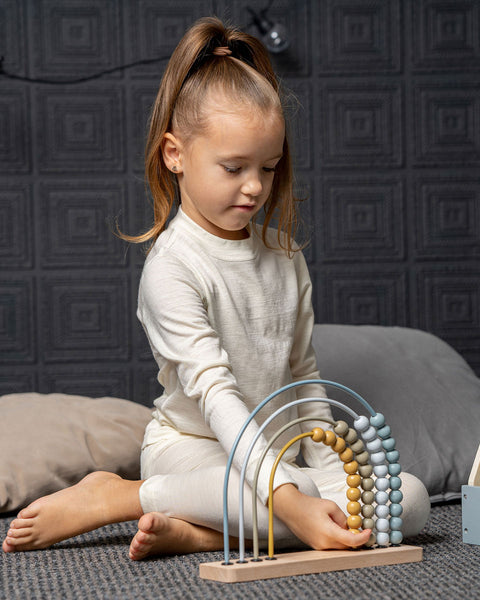 A young girl with her hair in a high ponytail sits cross-legged on a textured gray rug, wearing a cream-colored long-sleeve top and matching leggings. She is focused on playing with a wooden rainbow abacus toy, gently touching the colored beads.