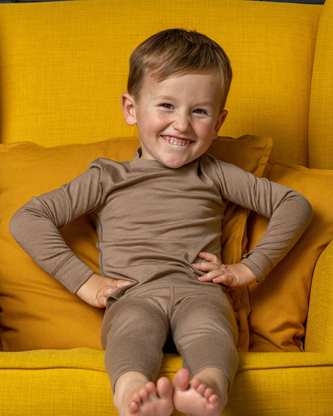 A young, barefoot boy sits playfully in a bright yellow armchair, wearing a matching taupe long-sleeve thermal top and leggings. He is smiling broadly at the camera with his hands placed confidently on his hips.