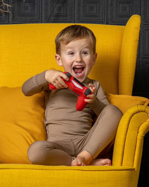 A joyful young boy wearing a taupe long-sleeve top and matching leggings sits comfortably in a vibrant yellow armchair. He is holding a bright red video game controller and laughing excitedly with his mouth wide open.