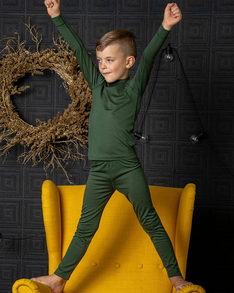 A young boy stands barefoot on a bright yellow armchair with his arms raised playfully in the air. He is wearing a dark green long-sleeve top and matching leggings, looking off to the side against a dark, textured wall decorated with a rustic wreath.