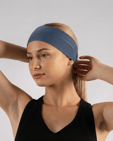 A fair-skinned woman with blonde hair pulled back wearing a black tank top and a denim-blue tie headband. She is looking slightly downward and to the side with her hands raised behind her head against a light gray background.