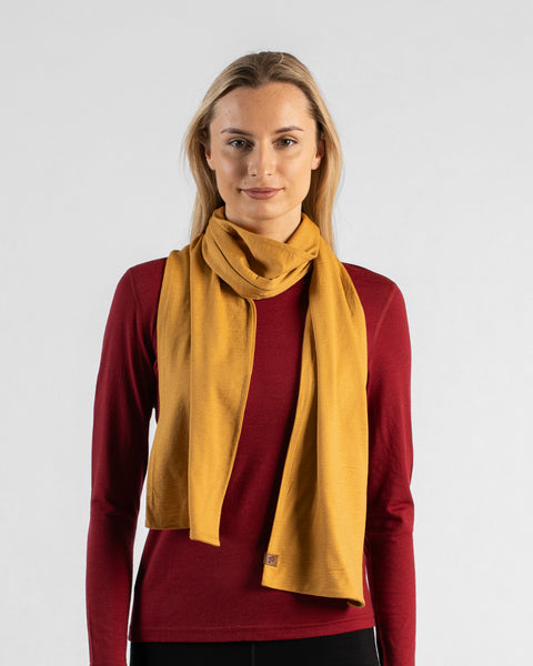 A studio portrait of a woman looking directly at the camera with a subtle smile. She is wearing a dark red long-sleeved shirt and a mustard yellow scarf wrapped once around her neck, with the ends hanging down in front. A small, square brown leather logo patch is visible on the bottom edge of the scarf. The background is a solid light grey.