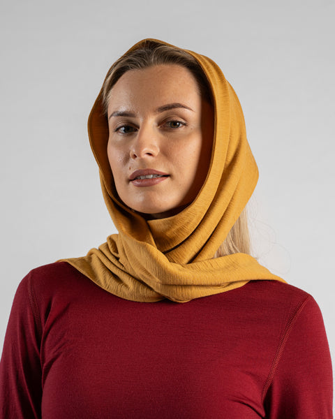 A close-up studio portrait of a blonde woman facing forward and looking directly at the camera against a plain light grey background. She is wearing a long-sleeved burgundy shirt and a mustard yellow scarf wrapped securely around her head and neck.