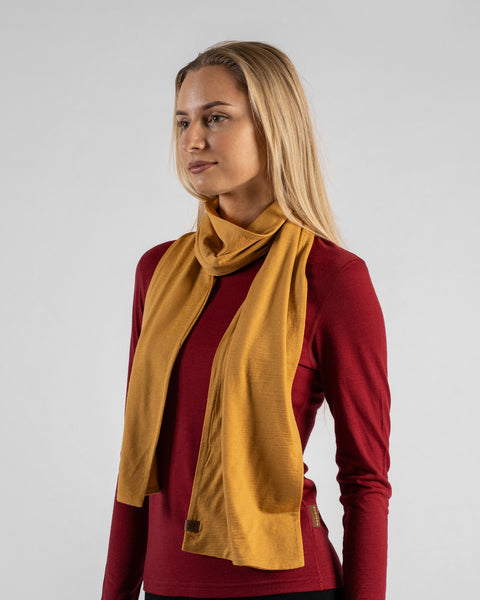 A studio portrait of a blonde woman shown in a three-quarter profile looking off to the left against a plain light grey background. She is wearing a long-sleeved burgundy shirt and a mustard yellow scarf draped loosely around her neck, with the ends hanging down in front.