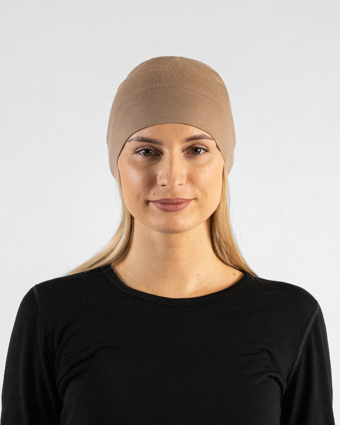 A studio portrait of a woman looking directly at the camera with a subtle smile. She is wearing a fitted, light beige skull cap over her blonde hair and a black top. The background is a solid light grey.