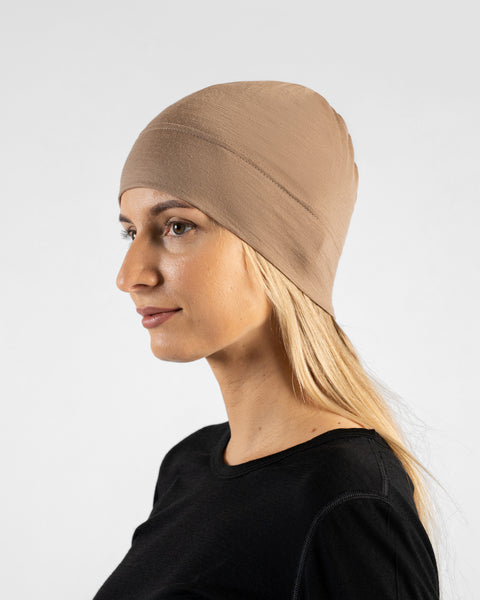 A studio portrait of a woman in a profile view, looking off to the left side of the frame. She is wearing a fitted, light beige skull cap over her blonde hair and a black top. The background is a solid light grey.