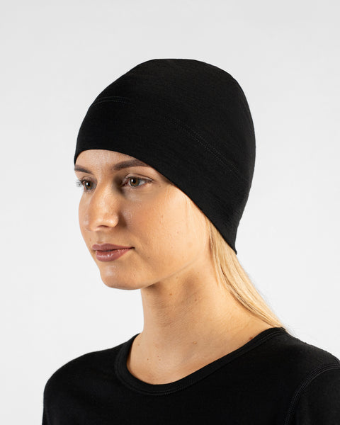 A studio portrait of a woman in a profile view, looking off to the left side of the frame. She is wearing a fitted, solid black skull cap over her blonde hair and a black top. The background is a solid light grey.