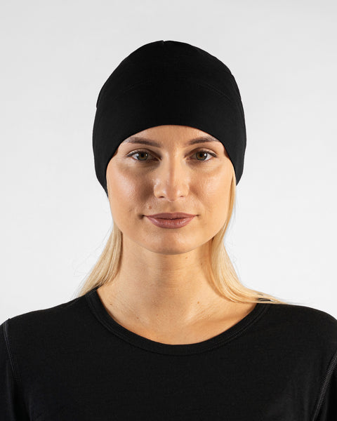 A studio portrait of a woman looking directly at the camera. She is wearing a fitted, solid black skull cap over her blonde hair and a black top. The background is a solid light grey.