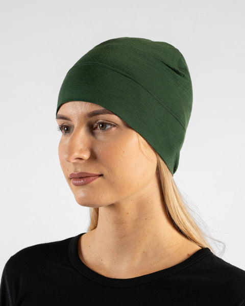 A studio portrait of a woman in a profile view, looking off to the left side of the frame. She is wearing a fitted, dark green skull cap over her blonde hair and a black top. The background is a solid light grey.