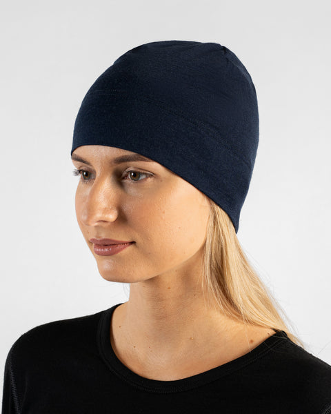 A studio portrait of a woman in a profile view, looking off to the left side of the frame. She is wearing a fitted, dark navy blue skull cap over her blonde hair and a black top. The background is a solid light grey.
