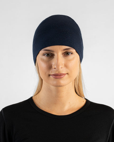 A studio portrait of a woman looking directly at the camera. She is wearing a fitted, dark navy blue skull cap over her blonde hair and a black top. The background is a solid light grey.