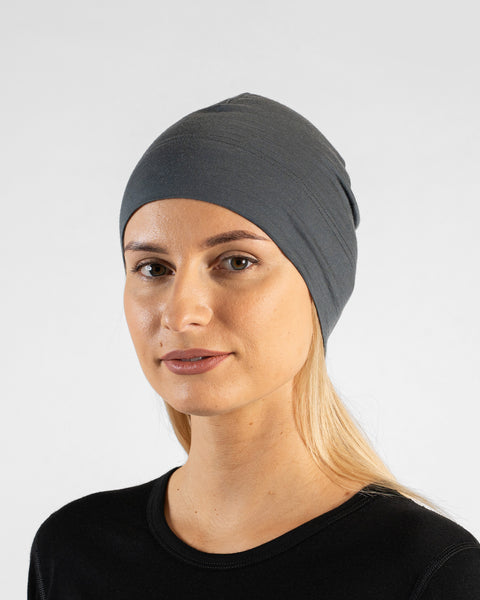 A studio portrait of a woman in a profile view, looking off to the left side of the frame. She is wearing a fitted, medium grey skull cap over her blonde hair and a black top. The background is a solid light grey.