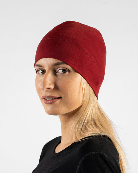 A studio portrait of a woman in a profile view, looking off to the left side of the frame. She is wearing a fitted, dark red (royal cherry) skull cap over her blonde hair and a black top. The background is a solid light grey.