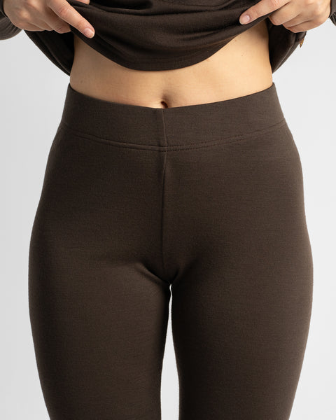 A close-up view of a woman slightly lifting her dark brown top to show the high-rise waistband and fit of matching brown thermal leggings.