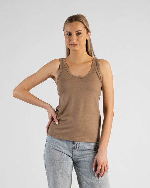 A studio portrait of a woman standing with her right hand resting on her hip, looking directly at the camera. She is wearing a fitted, beige scoop-neck sleeveless tank top and light-wash blue jeans. The background is a solid, light grey.