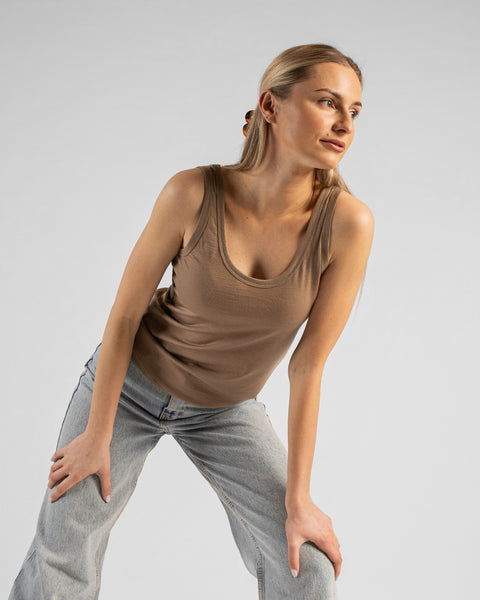 A studio portrait of a woman leaning slightly forward with her hands resting on her thighs, looking off to the right side of the frame. She is wearing a fitted, beige scoop-neck sleeveless tank top and light-wash blue jeans. Her blonde hair is partially pulled back with a claw clip. The background is a solid, light grey.