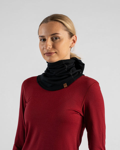 A studio portrait of a blonde woman wearing a burgundy long-sleeved shirt and a black balaclava pushed down around her neck like a thick collar, against a plain light grey background.