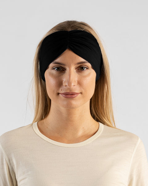 A close-up studio portrait of a blonde woman facing forward, looking directly at the camera with a soft smile against a plain light grey background. She is wearing a light beige long-sleeved shirt and a black ribbon headband.