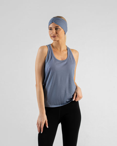 A studio portrait of a blonde woman standing and looking slightly off to her left. She is wearing a dusty blue sleeveless top, black leggings, and a matching wide dusty blue headband with a gathered twist detail at the front, against a plain light grey background.