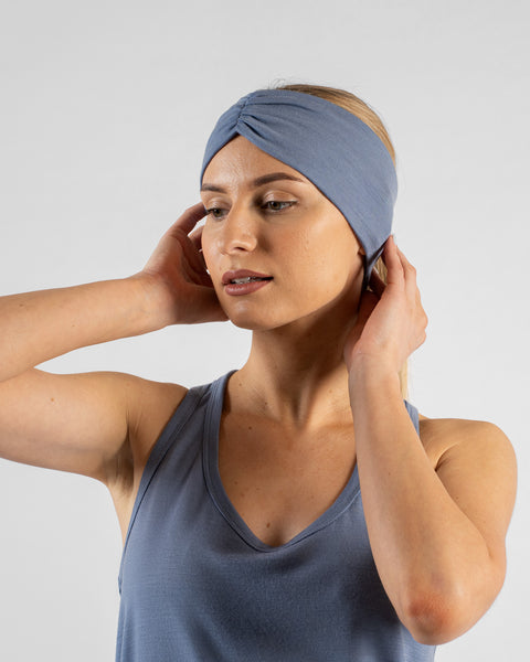 A close-up studio portrait of a blonde woman looking downward and slightly to her left, with her hands gently adjusting the sides of a wide dusty blue headband with a gathered twist detail at the front. She is wearing a matching dusty blue sleeveless top against a plain light grey background.