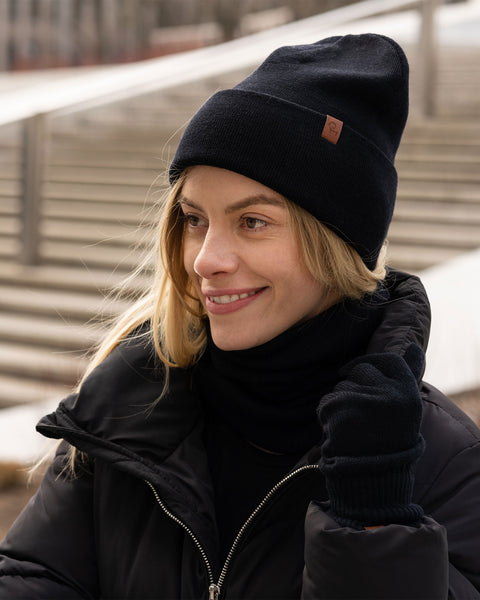 A smiling woman standing outdoors, looking happily off to the side. She is bundled up in a black puffer jacket and a matching set of black knit accessories: a beanie with a brown leather logo patch, a thick neck gaiter, and gloves. Light-colored concrete steps are visible in the soft-focus background.