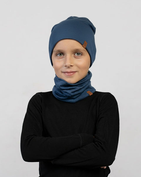 A child wearing a dark blue beanie and neck gaiter set with a black long-sleeve shirt poses with arms crossed and a slight smile against a light gray background.