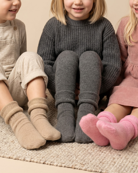 A cropped shot showing the lower halves of three young children sitting side-by-side on a textured rug. They are modeling thick, ribbed knit socks folded down at the ankles. The child on the left wears beige socks with beige overalls; the middle child wears dark grey socks with matching grey ribbed tights and a sweater; the right child wears pink socks with a pink corduroy dress.