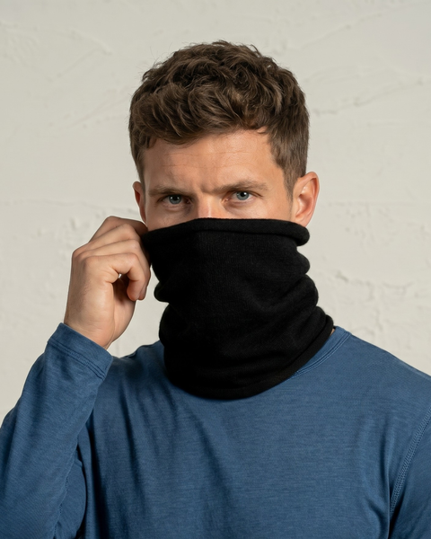 A man looking directly at the camera, wearing a blue long-sleeved shirt and a black knitted neck gaiter pulled up to cover his mouth and nose. His right hand is holding the top edge of the gaiter. The background is a textured off-white wall.