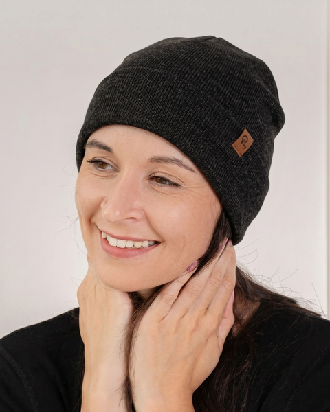 A close-up portrait of a smiling woman looking to the side. She is wearing a black knitted beanie featuring a small square brown leather logo patch, paired with a black long-sleeved top. Her hands rest gently near her neck. The background is a white paneled wall.