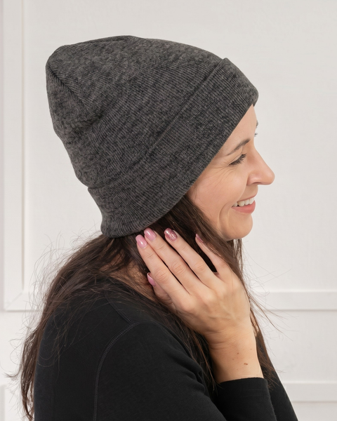 A close-up profile shot of a smiling woman looking to the right. She is wearing a dark grey heathered knit beanie and a black long-sleeved top. One hand rests gently on her dark hair near her neck. The background is a bright white wall with subtle paneling.