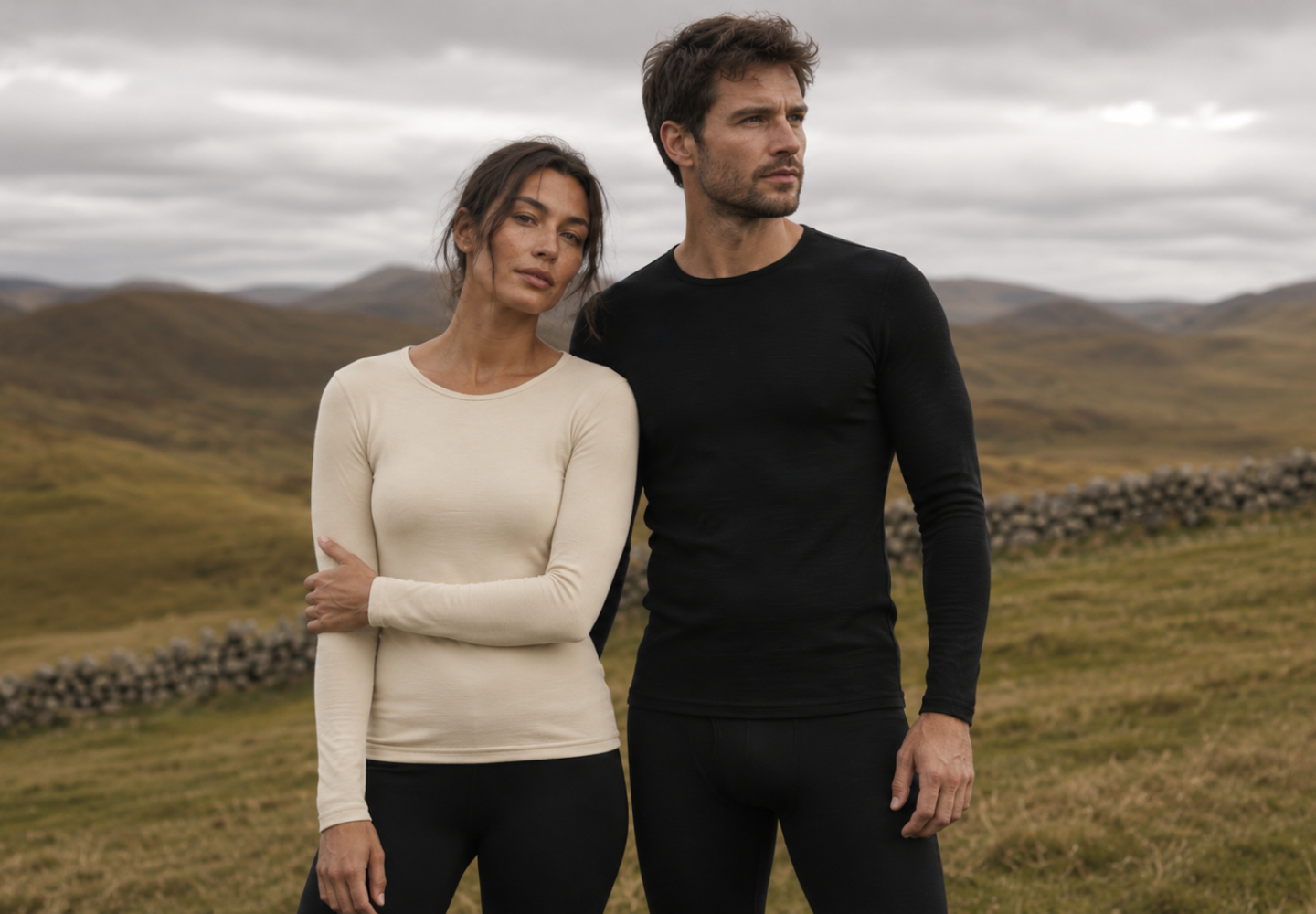 A woman and a man stand side-by-side in a rugged, grassy landscape under a cloudy gray sky. The woman is wearing a cream-colored long-sleeved thermal top while the man wears a matching black thermal set, both looking thoughtfully off-camera.