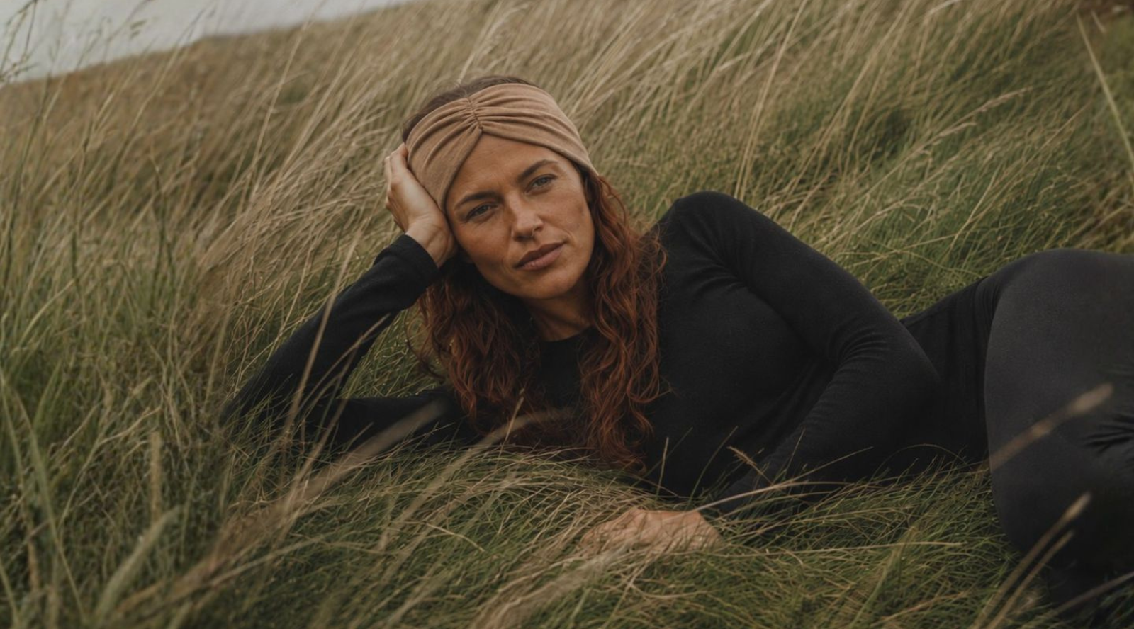 A woman with long auburn hair reclines in tall, dry grass while wearing a black long-sleeved top and leggings, accessorized with a tan twisted headband. She rests her head on her hand, looking thoughtfully toward the camera.