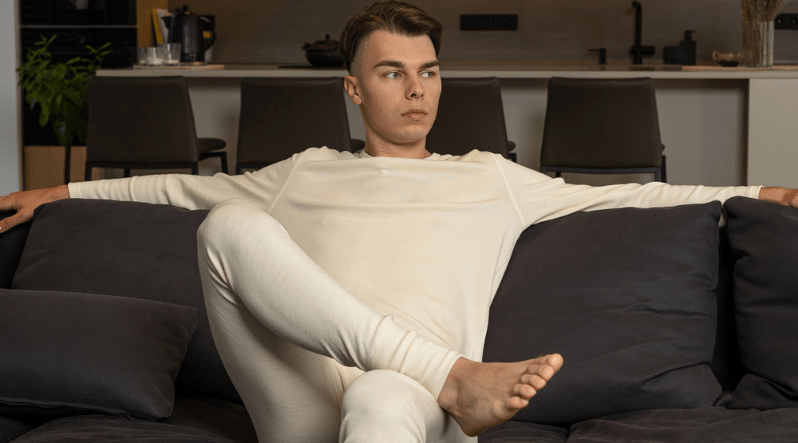 a man sitting on a couch, wearing the merino wool matching set in natural color, consisting of a long sleeve shirt and pants.