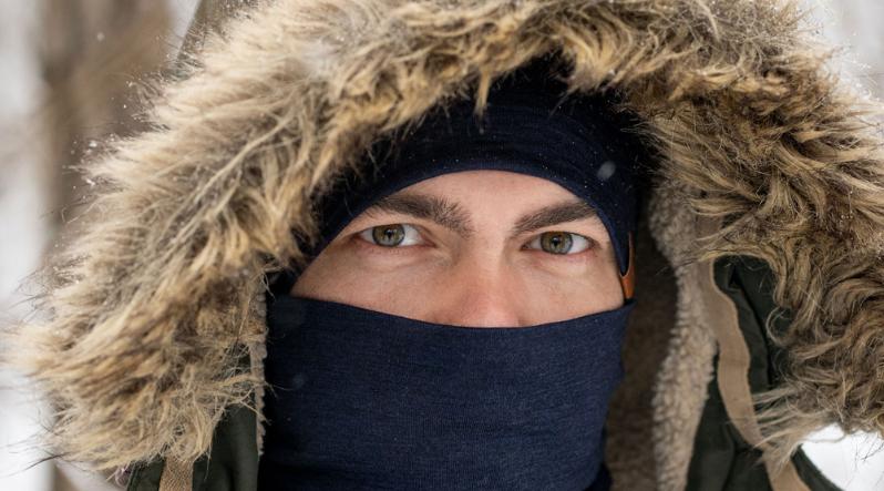 A man wearing the hood of a winter jacket over his head, with a denim color merino wool neck gaiter covering his face up to the nose bridge.