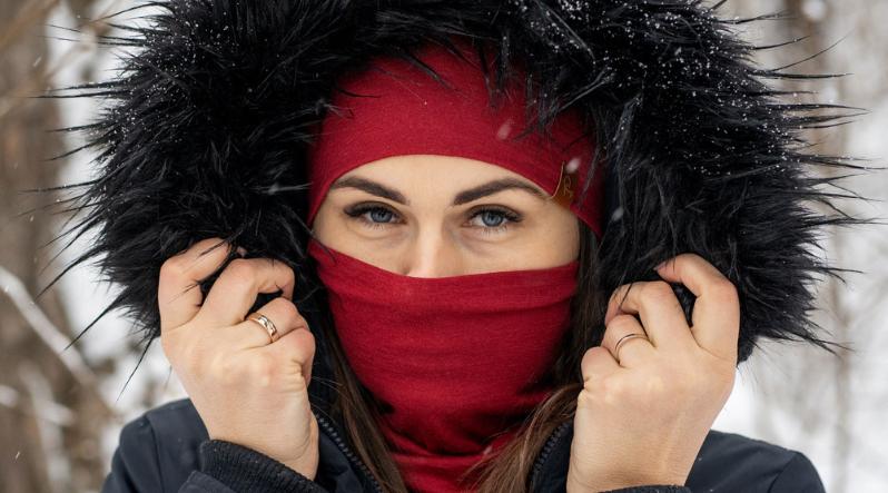 A woman standing in a snowy forest, with her winter jacket hood on, holding twh sides of it with her hands. She is wearing royal cherry neck gaiter and a matching beanie.
