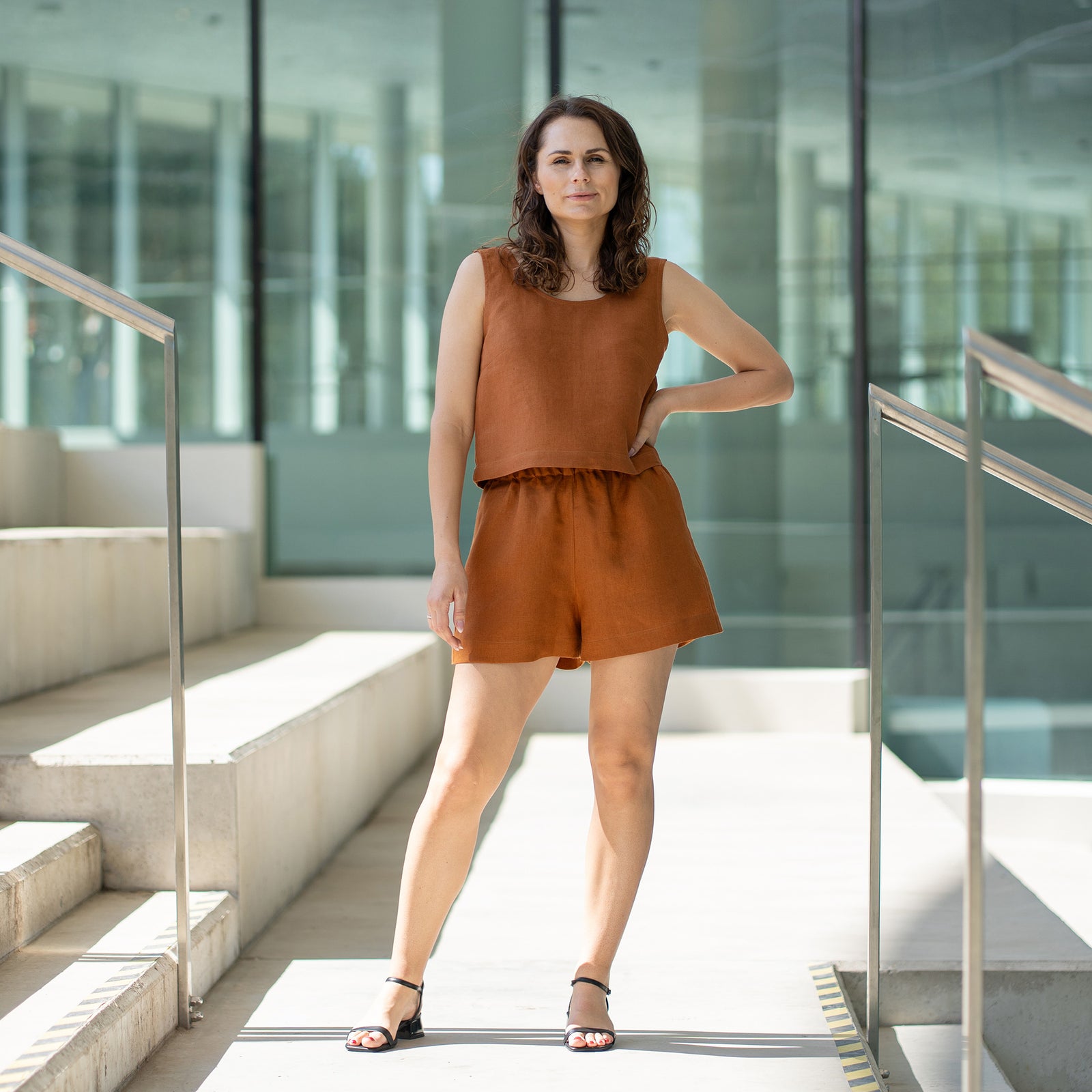 A woman with brown hair, wearing a matching almond brown sleeveless top and shorts, looks over her shoulder while standing on outdoor concrete stairs with metal railings.