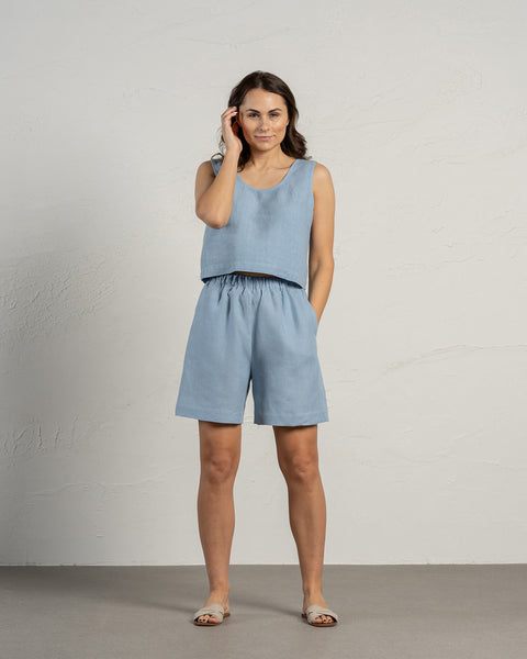 A woman with dark hair wearing 100% linen shorts in the color cloudy blue and linen top in the color cloudy blue. Her one hand is in the pocket while the other is touching her hair. The background of the photo is in natural, nude colors.