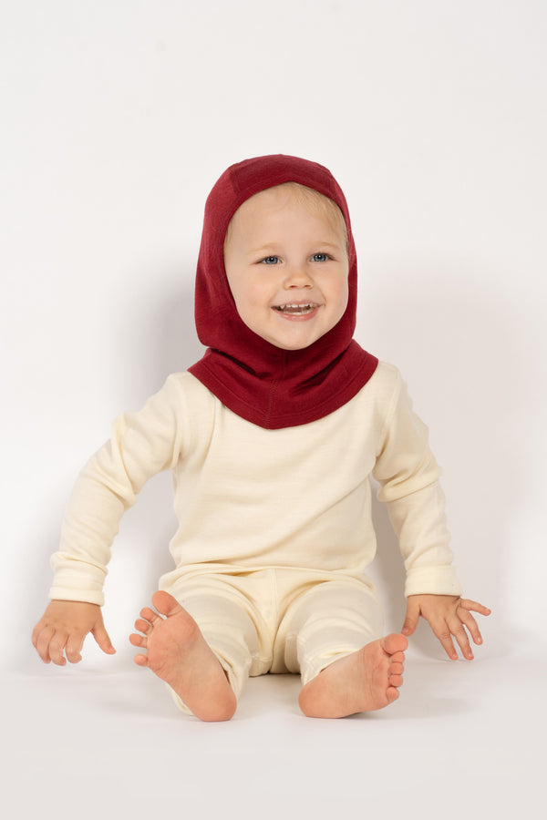 A toddler girl sitting on the ground, wearing the menique kids' merino 160gsm balaclava in royal cherry color, studio background picture.