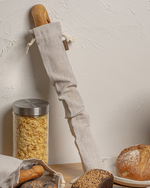 A crusty baguette partially enclosed in a long, natural-colored linen drawstring bag, leaning vertically against a textured white wall. It sits on a wooden counter next to a glass storage jar filled with cereal, a sliced bagel in a small linen bag, and a round loaf of artisan bread.