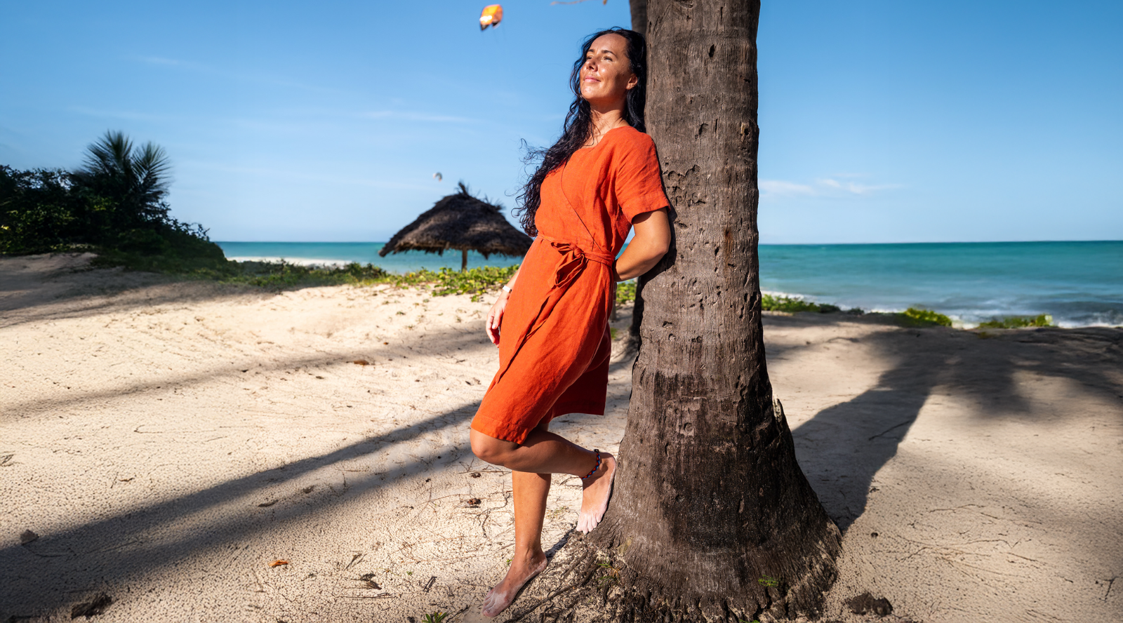 A woman with long dark hair relaxing on a sunny beach, leaning her back against a palm tree trunk with her eyes closed. She is barefoot and wearing a short-sleeved, rust-orange linen wrap dress tied at the waist. The background features white sand, turquoise ocean water, a thatched sunshade, and a kite surfer in the distance under a clear blue sky.