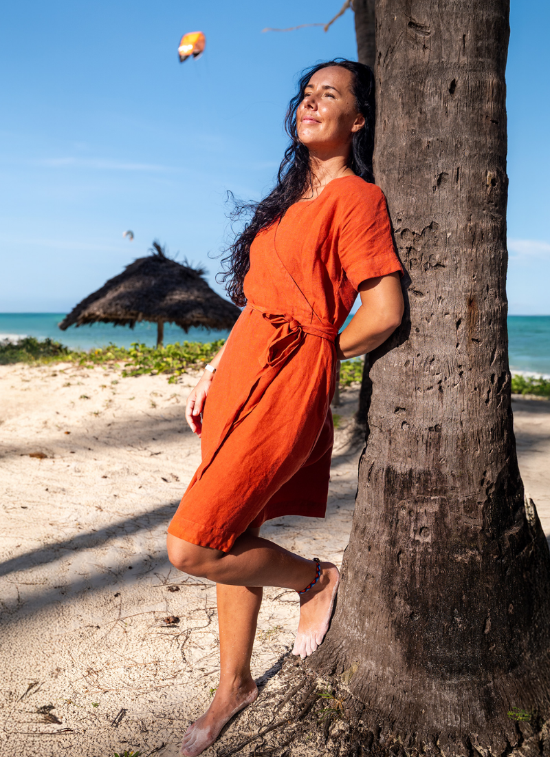 A woman with long dark hair relaxing on a sunny beach, leaning her back against a palm tree trunk with her eyes closed. She is barefoot and wearing a short-sleeved, rust-orange linen wrap dress tied at the waist. The background features white sand, turquoise ocean water, a thatched sunshade, and a kite surfer in the distance under a clear blue sky.