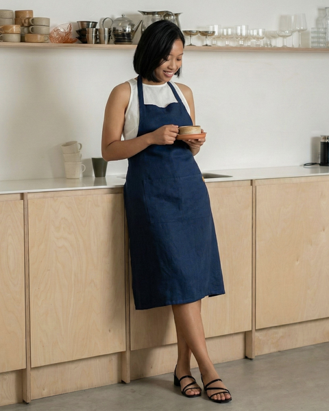 Woman wearing a storm blue linen bib apron, standing in a modern kitchen and holding a cup of coffee.