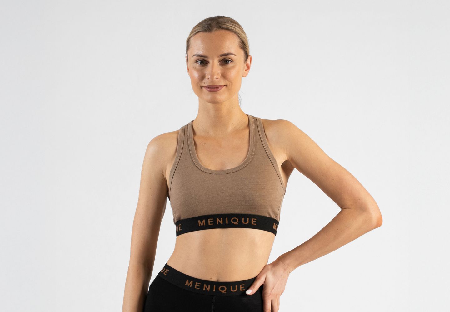A studio portrait of a smiling blonde woman standing with her left hand resting on her hip, looking directly at the camera. She is wearing a light brown or beige sports bralette featuring a black elastic underbust band with "MENIQUE" woven in light brown text. The top edge of a matching black "MENIQUE" waistband from her bottoms is visible below. The background is a solid light grey.