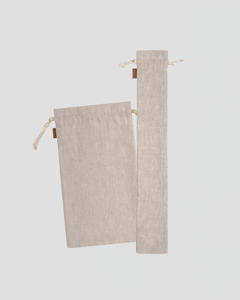 A studio laydown of two natural beige linen bread bags against a plain light gray background. The set includes one medium rectangular bag and one long, slender baguette bag, both featuring natural rope drawstrings and small brown brand tags.