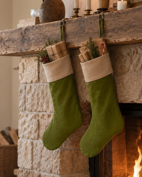 Two green Christmas stockings with beige cuffs hang from a rustic wooden mantel above a stone fireplace. The stockings are filled with wrapped gifts and holiday greenery, with candles and decor visible on the mantel and a fire burning in the fireplace below.