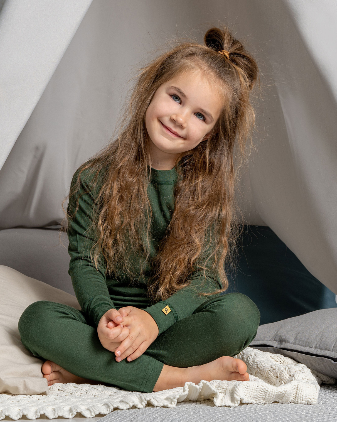 A young girl with long, curly brown hair tied in a high bun sits cross-legged inside a white fabric teepee. She is smiling and wearing a dark green, ribbed long-sleeved top and matching leggings.