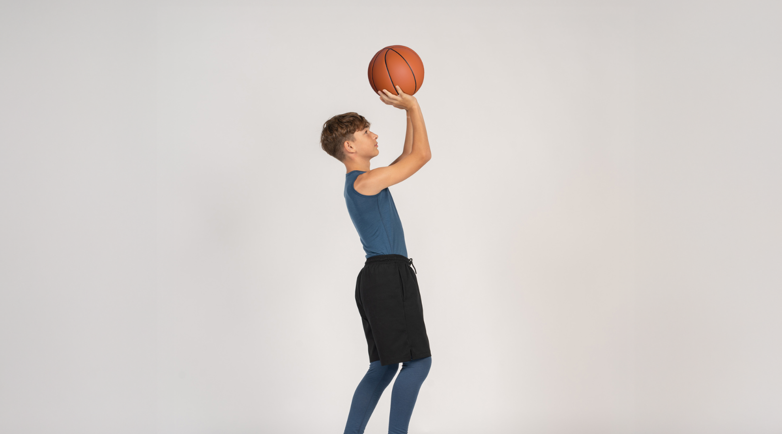 A young boy in athletic wear prepares to shoot a basketball against a plain light grey studio background. He is wearing a blue tank top, black shorts layered over blue leggings, and white sneakers.