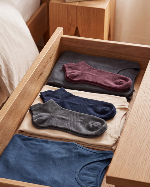 An open light wood drawer filled with neatly folded dark grey, beige, and blue t-shirts. Three pairs of ankle socks in burgundy, navy blue, and grey are laid out flat on top of the shirts, with a bed and wooden nightstand partially visible in the background.