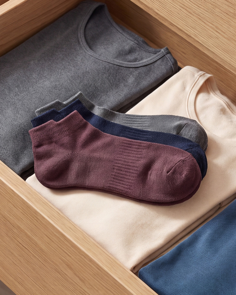 A close-up of an open light wood drawer containing folded dark grey, beige, and blue t-shirts. Three pairs of ankle socks in grey, navy blue, and burgundy are stacked neatly together on top of the beige shirt.